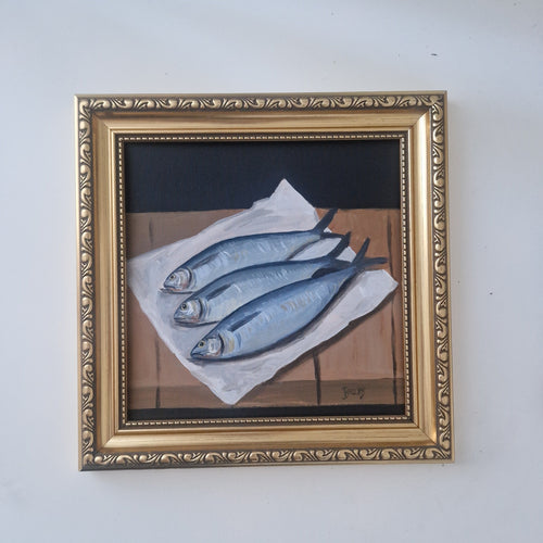 A framed still life painting of three blue-gray fish on white paper, set in an ornate gold frame and hung on a plain white wall.