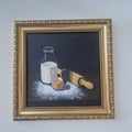 A framed still life painting on a dark background, depicting a glass bottle of milk, a cracked egg, and a rolling pin, surrounded by flour. The artwork is housed in an ornate gold-colored frame and is mounted on a plain light gray wall.