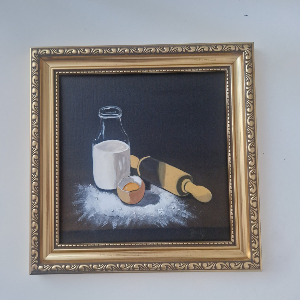 A framed still life painting on a dark background, depicting a glass bottle of milk, a cracked egg, and a rolling pin, surrounded by flour. The artwork is housed in an ornate gold-colored frame and is mounted on a plain light gray wall.