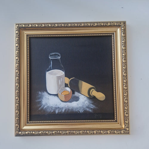 A framed still life painting on a dark background, depicting a glass bottle of milk, a cracked egg, and a rolling pin, surrounded by flour. The artwork is housed in an ornate gold-colored frame and is mounted on a plain light gray wall.